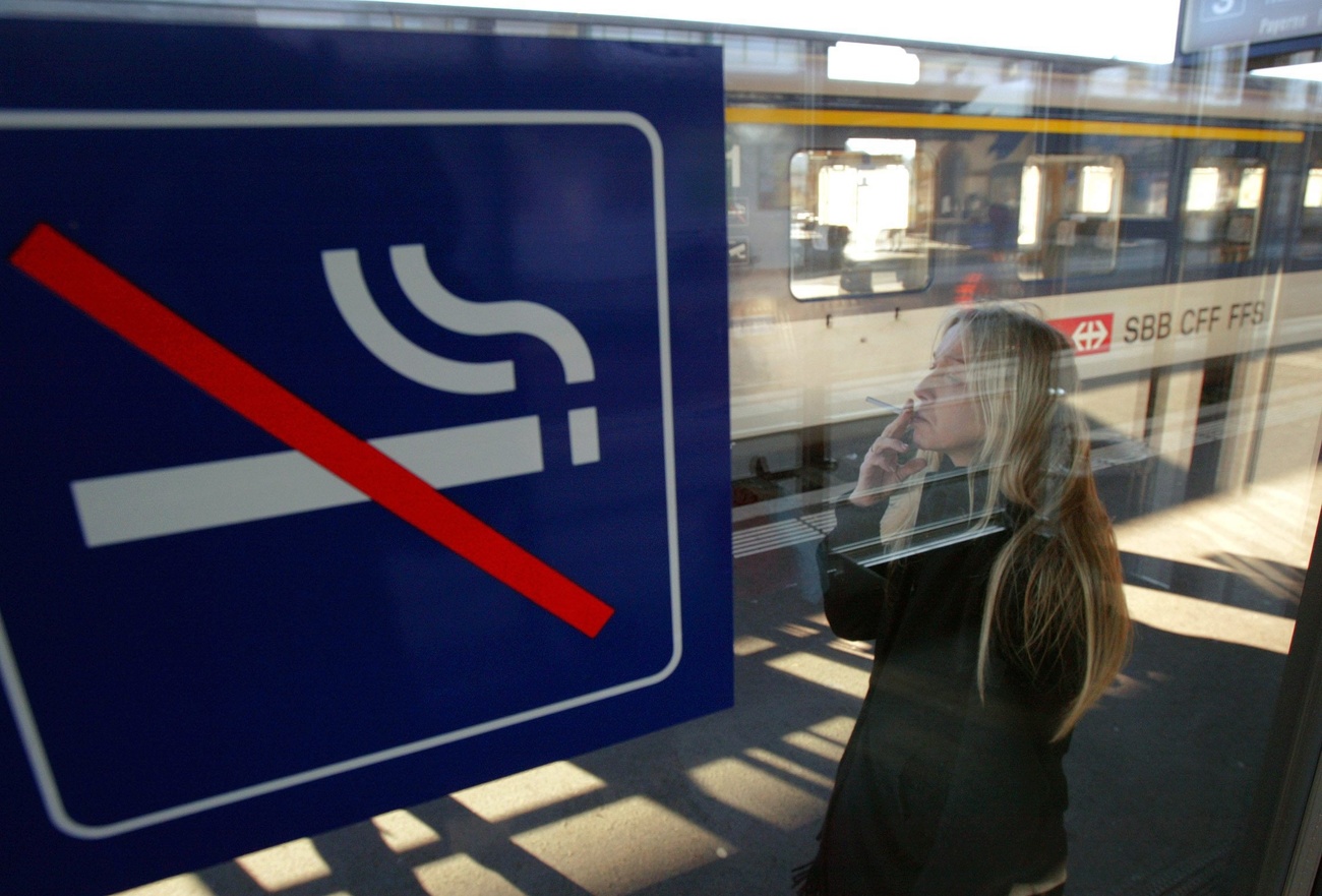 Mulher fuma cigarro em uma estação de trem suíça ao lado de um sinal de não fumantes
