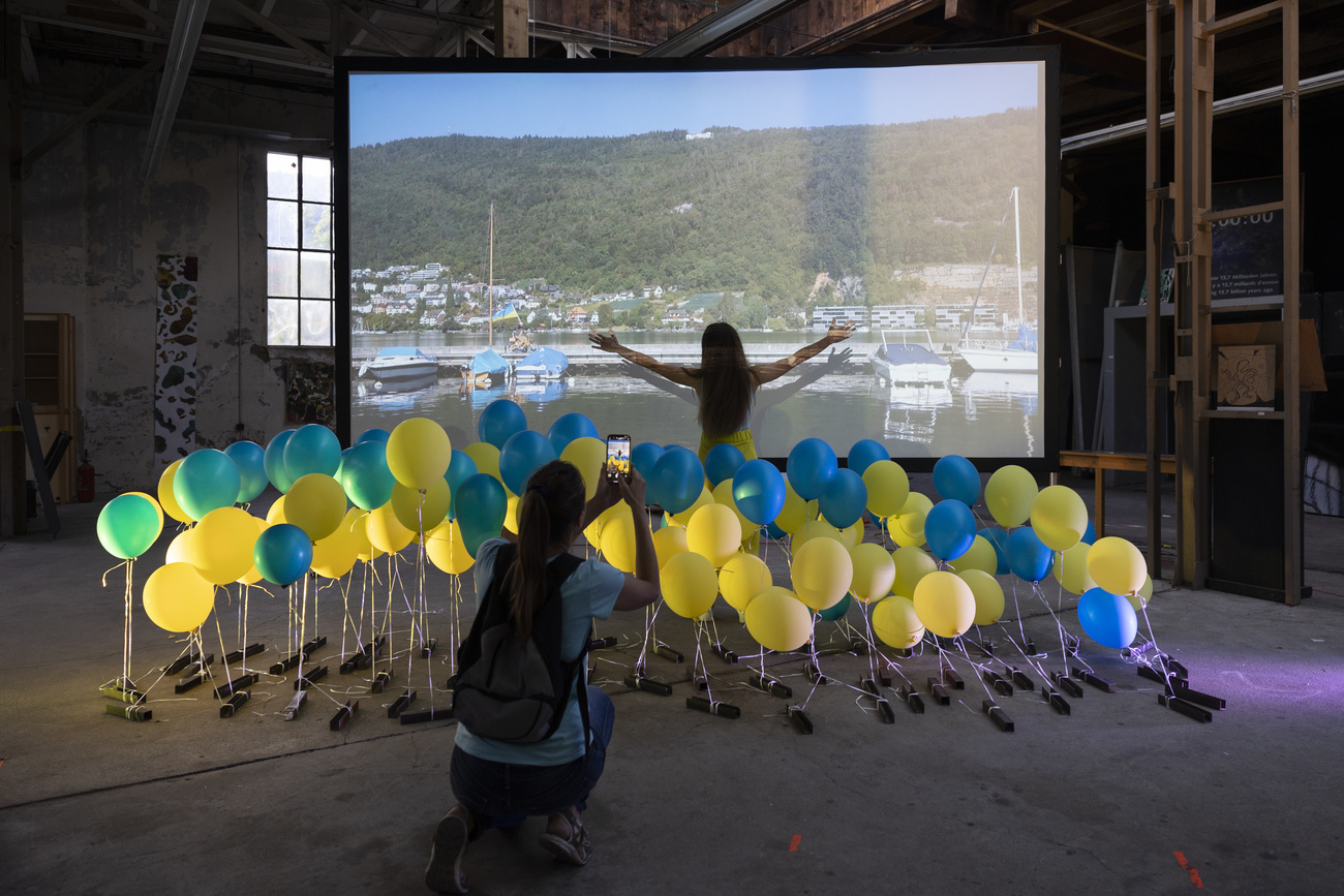 Una mujer fotografía globos azules y amarillos y otra mujer abre los brazos delante de una pantalla