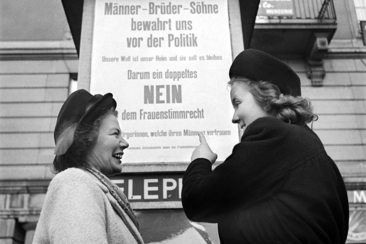 Foto en blanco y negro. Dos mujeres señalando un cartel de publicidad contra el voto de la mujer