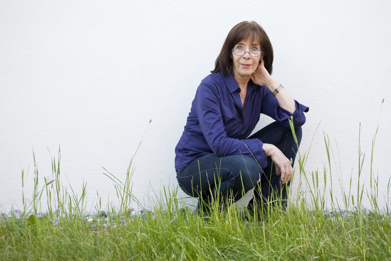 donna con capellis curi e occhiali in jeans e camicia blu accovacciata su un prato erboso dietro di lei un muro bianco