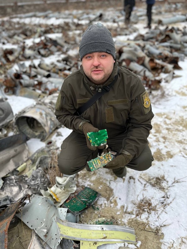 Mann in Militärkleidung in der Hocke auf einem Feld mit Materialtrümmern