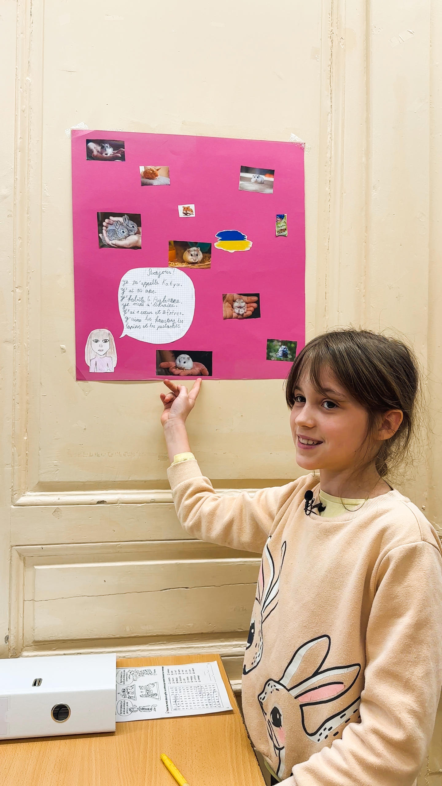 Девочка показывает украшенную ею доску с изображениями хомячков, украинского флага и рассказа о себе на французском языке