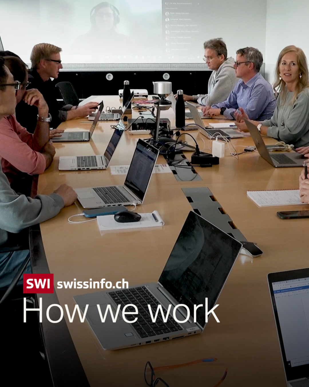 Picture shows a long table with people around it, discussing something, and someone also on via a video call in the background.