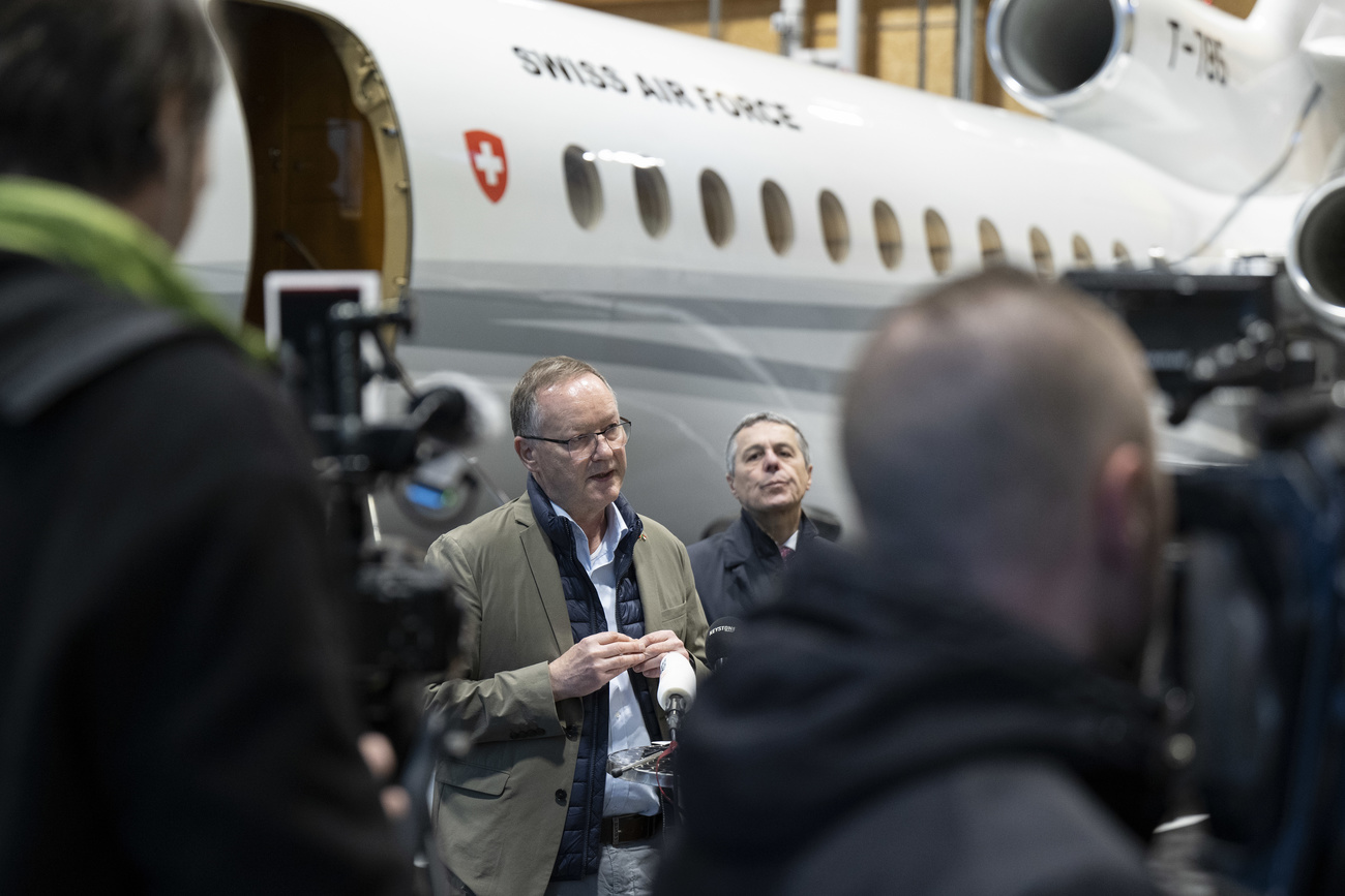 two men stand in front of a plane speaking to journalists