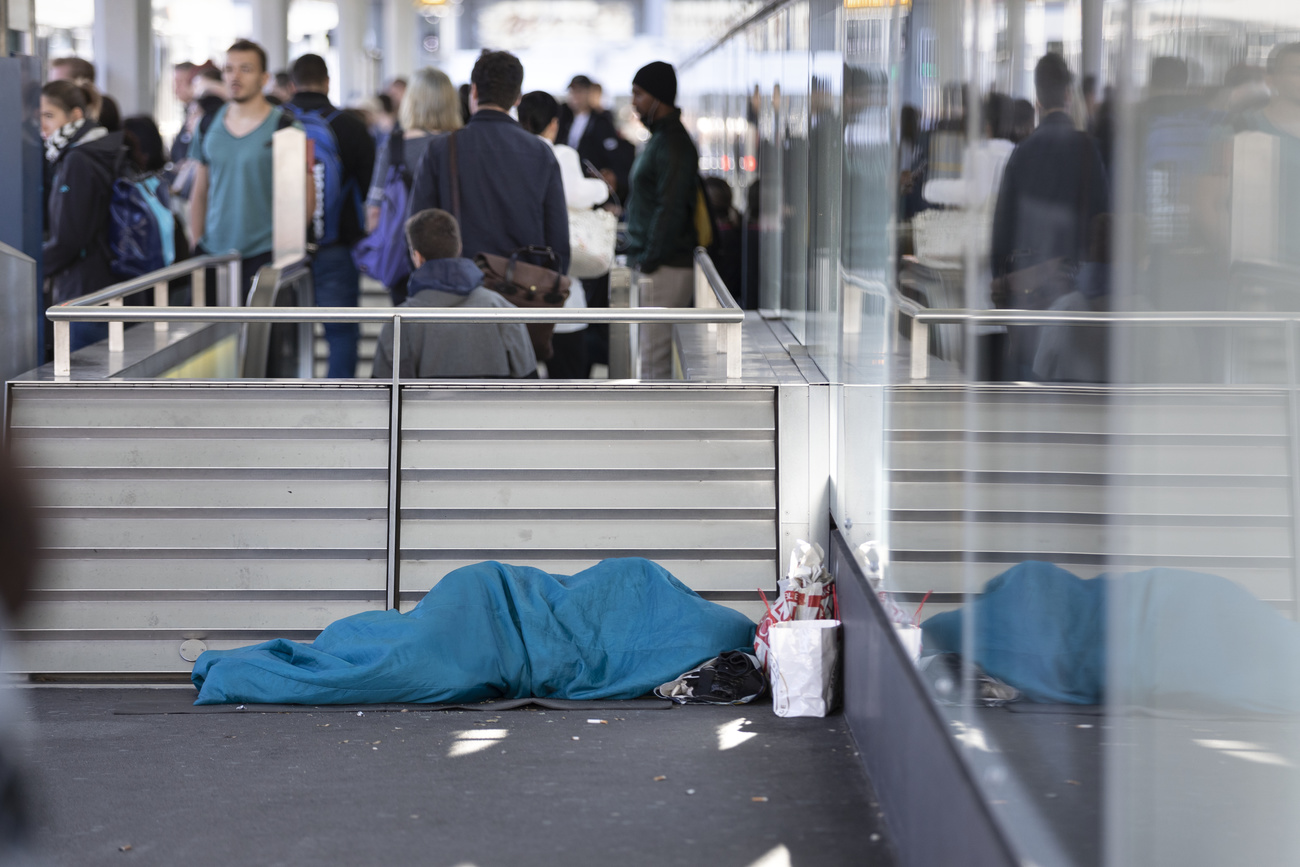 Person, die am Bahnhof Zürich in einem blauen Schlafsack inmitten von Pendler:innen am Boden schläft