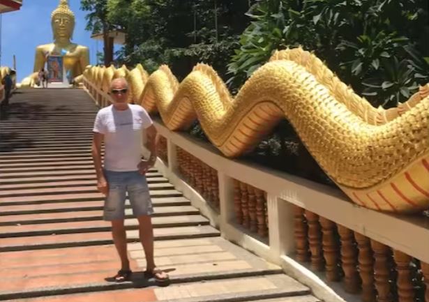 A swiss man living abroad in Thailand stands by a gold dragon handrail leading up to a gold buddha.