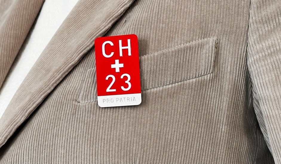 Retor-Velo-Plakette als 1. August Abzeichen. Hintergrund: Rot. Oben: CH. Mitte: Schweizrkreuz. Unten: 23.