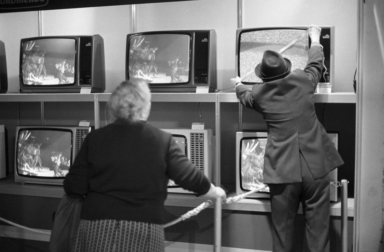 black-and-white photo of television sets, and a man measures one with measuring tape
