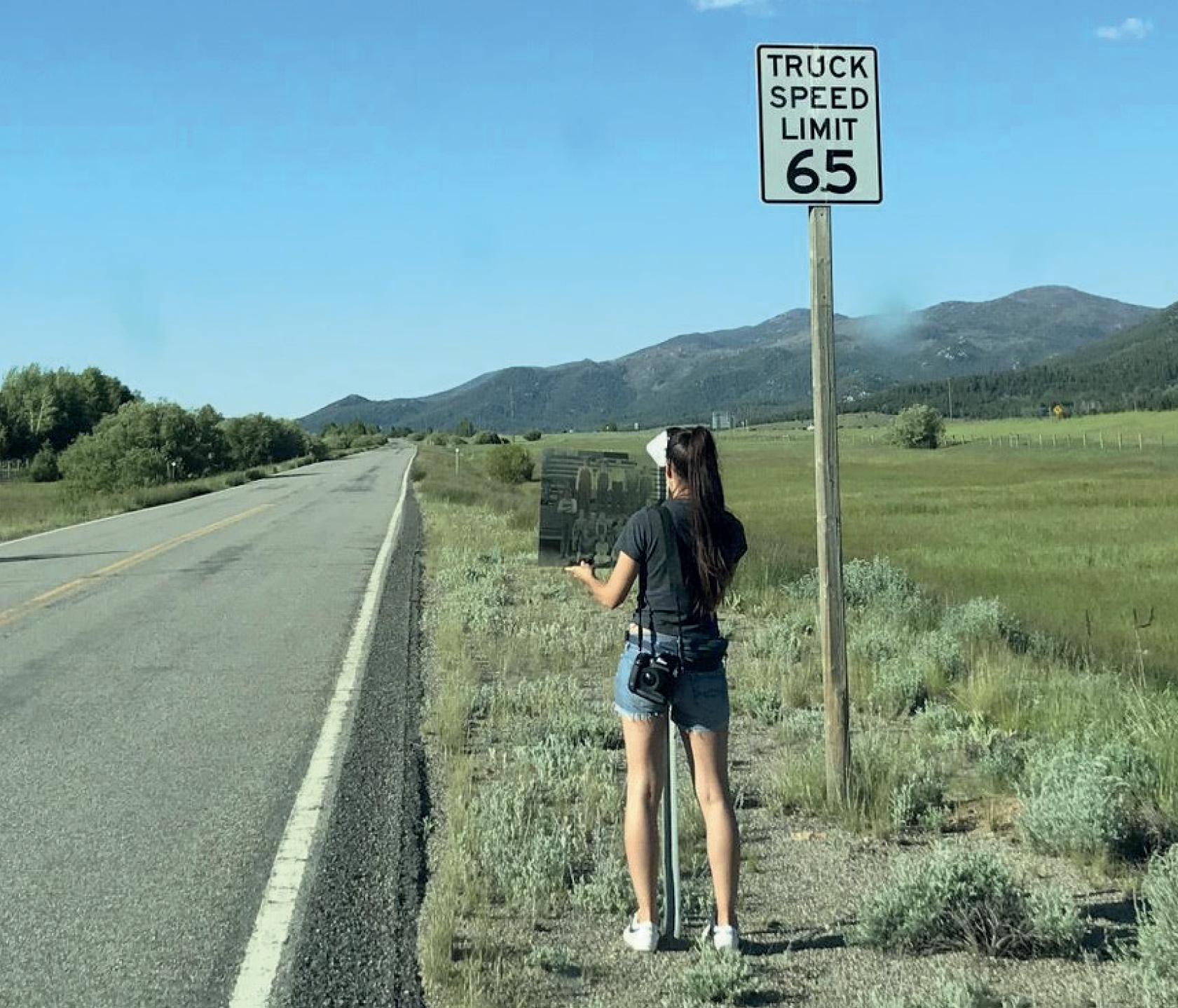 A woman holding a photograph at a roadside- where a road sign reads: Truck Speed Limit 65