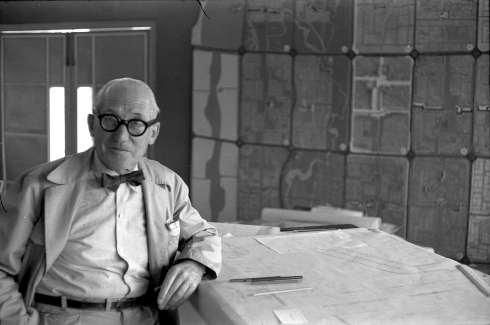Der Architekt Le Corbusier schaut direkt in die Kamera und stützt sich mit dem Ellenbogen auf einen Entwurf