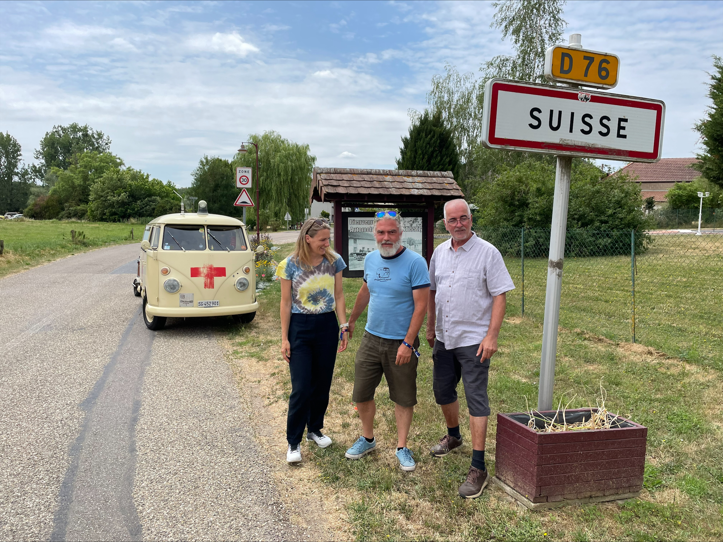 Eine Frau und zwei Männer vor einem Ortsschild mit Suisse drauf.