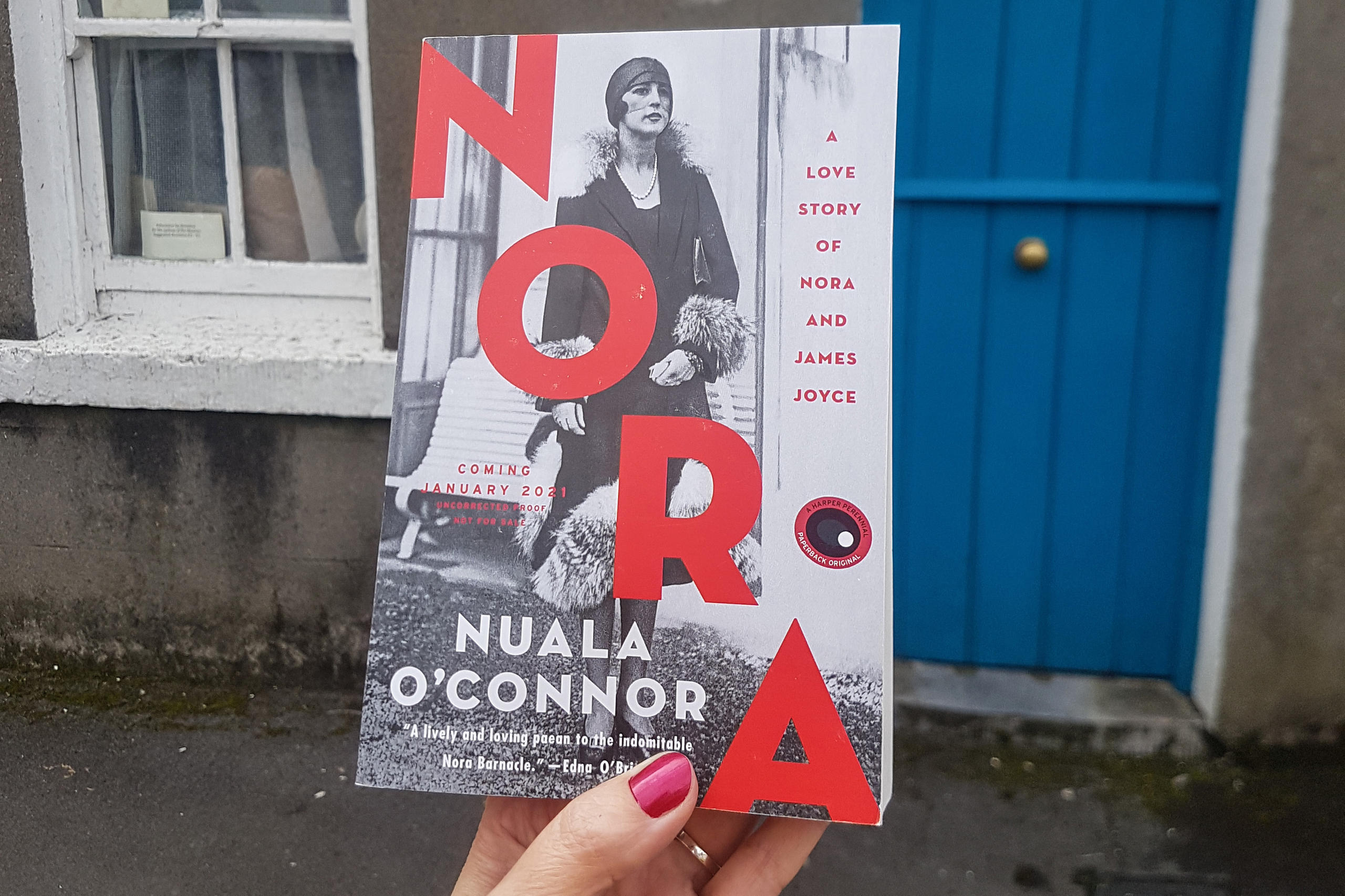 A hand holding a copy of the book Nora in front of Nora Joyce s house in Galway