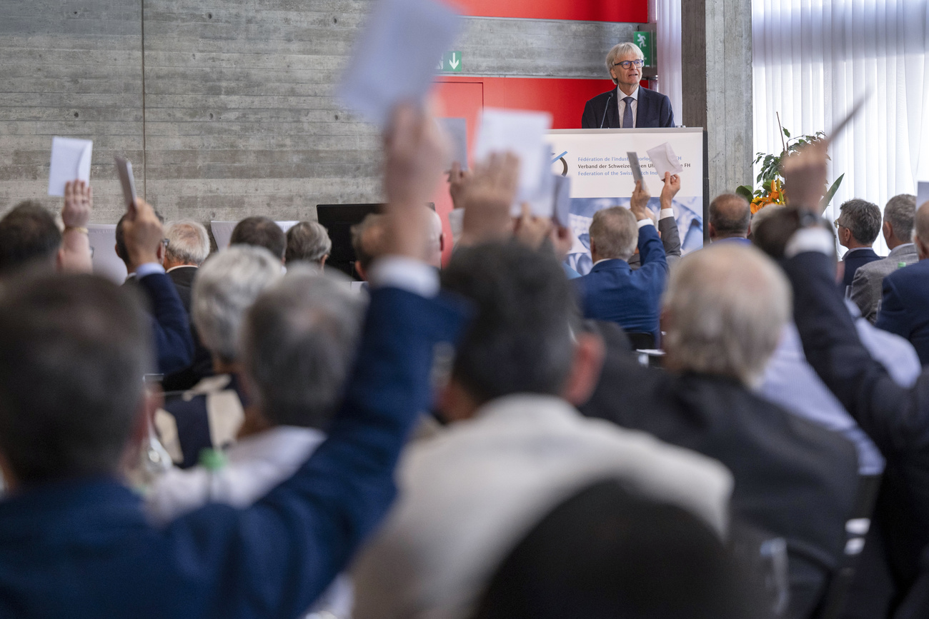 Jean-Daniel Pasche auf einem Podium, im Saal wird abgestimmt, einige der Besucher haben dazu die Hand erhoben