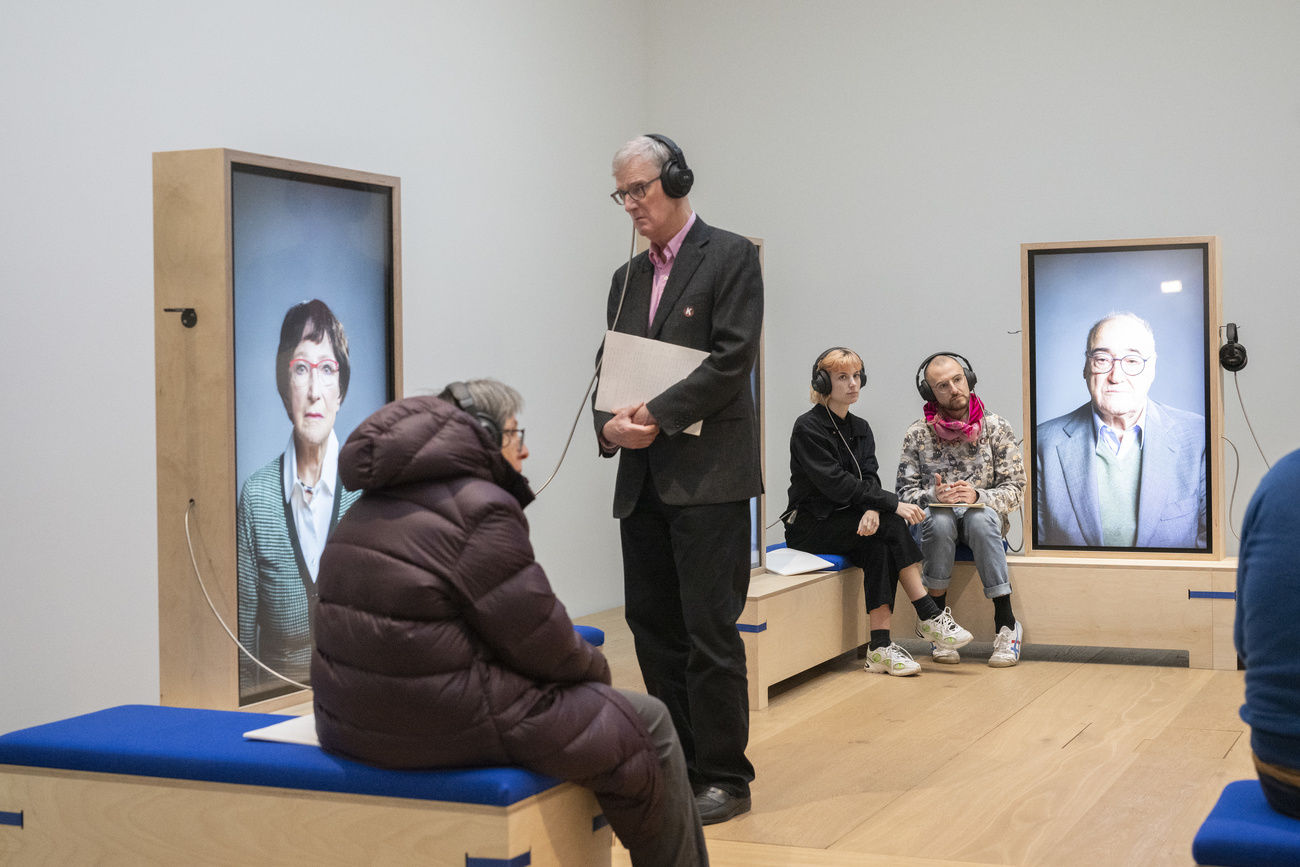 Museumsbesucher:innen in einem Saal mit Bänken, Kopfhörern und hinterleuchteten Kästen mit den Portraits von Persönlichkeiten