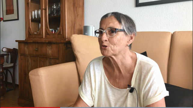 Eine ältere Frau sitzt auf einem hellbraunen Sofa und blickt in die Kamera.