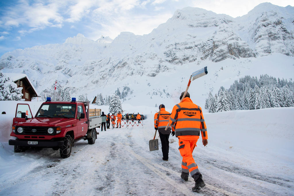 Uomini con tuta arancione attrezzati di pala camminano verso un mezzo di soccorso e una barriera in un paesaggio innevato