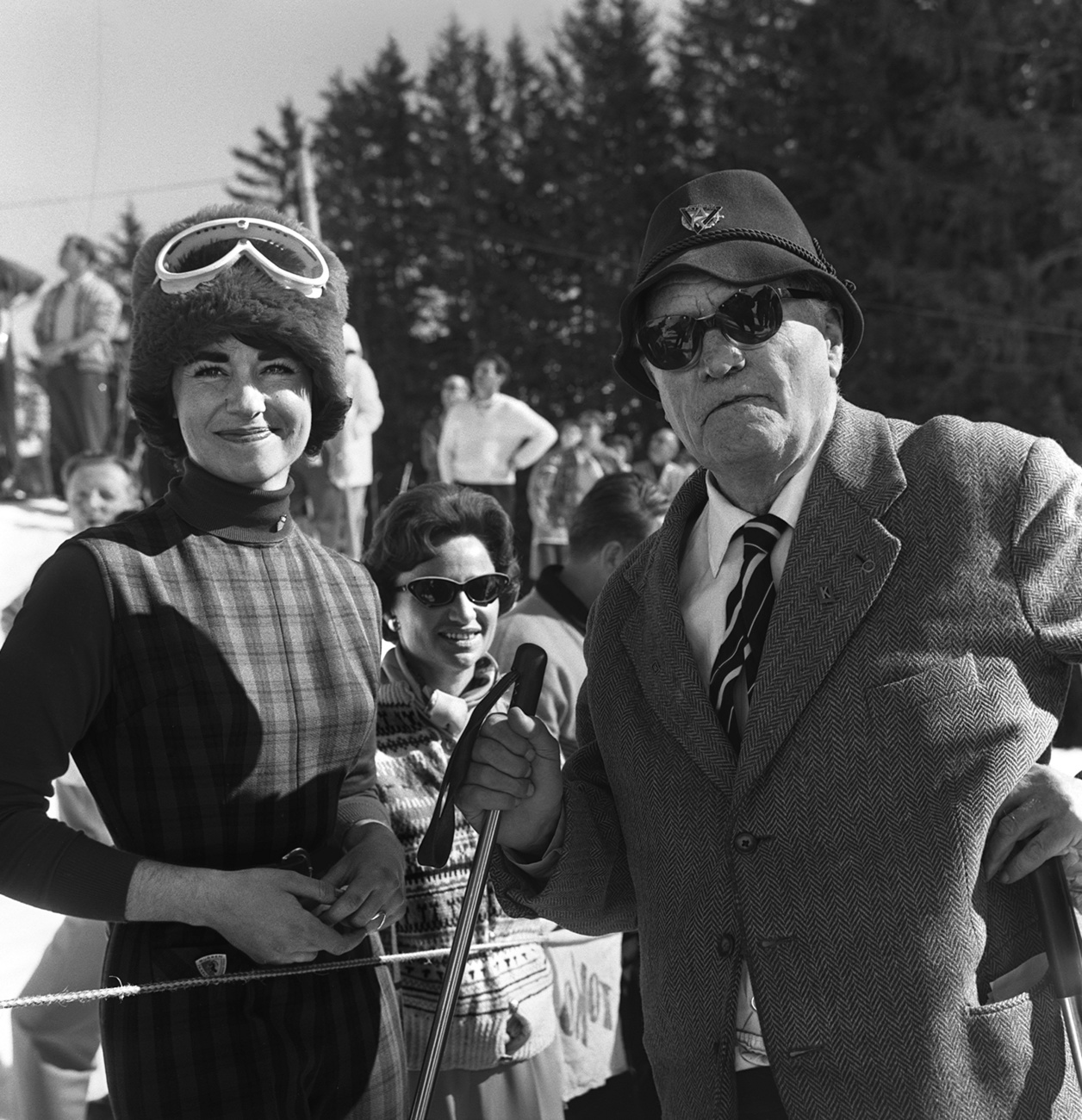 Arnold Lunn posiert im Anzug mit Skistöcken neben einer jungen Frau auf der Piste für die Kamera