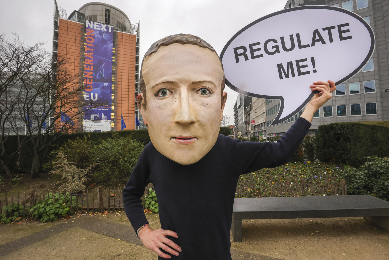 persona con maschera di cartapesta rappresentante mark zuckerberg tiene cartello con scritta REGULATE ME!