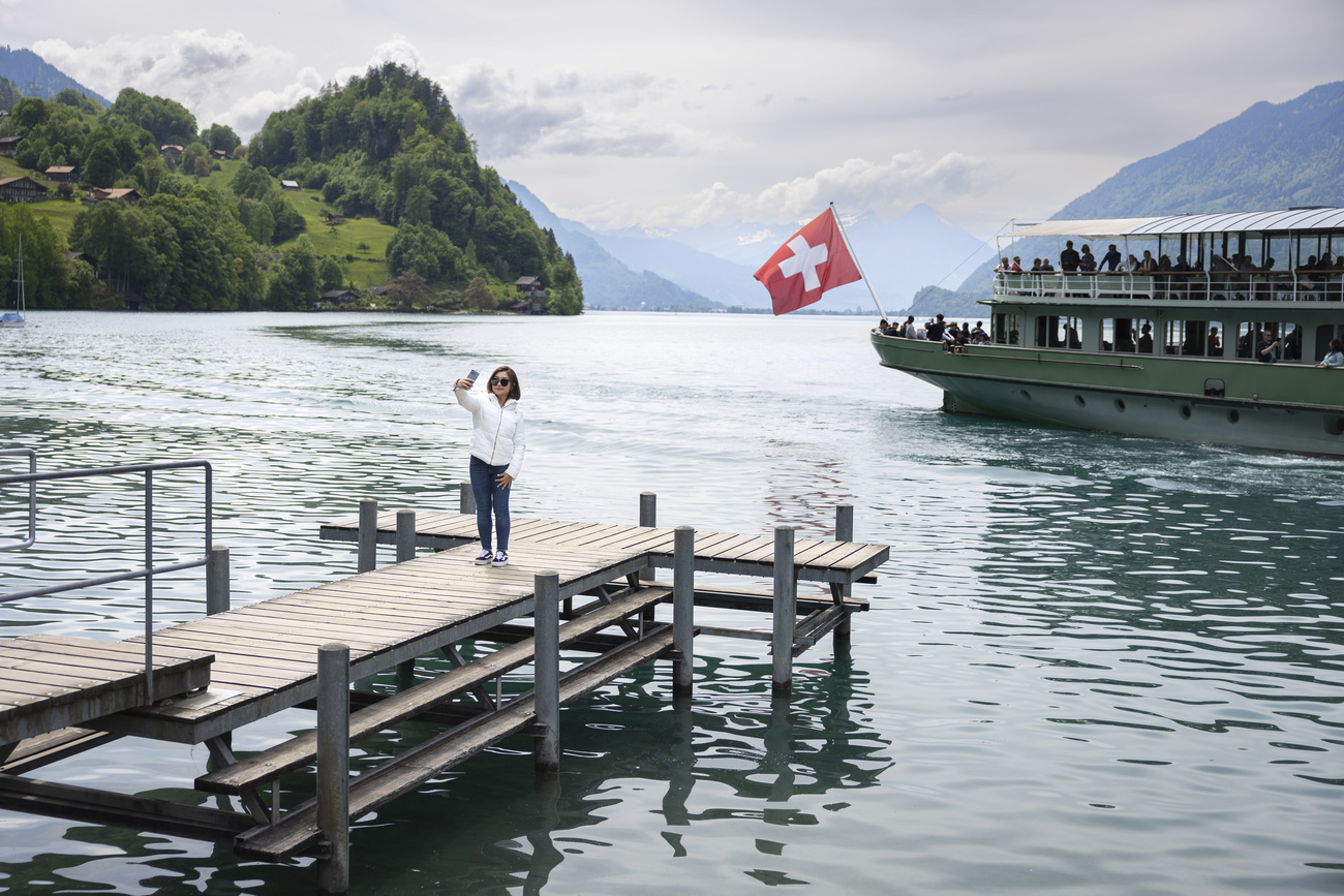 turista si scatta un selfie su un molo mentre dietro di lei passa un battello che batte bandiera svizzera