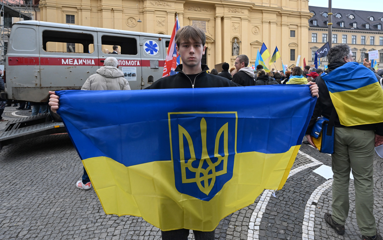 Ein Demonstrant mit einer Ukraine-Fahne vor einem zerschossenen Krankenwagen