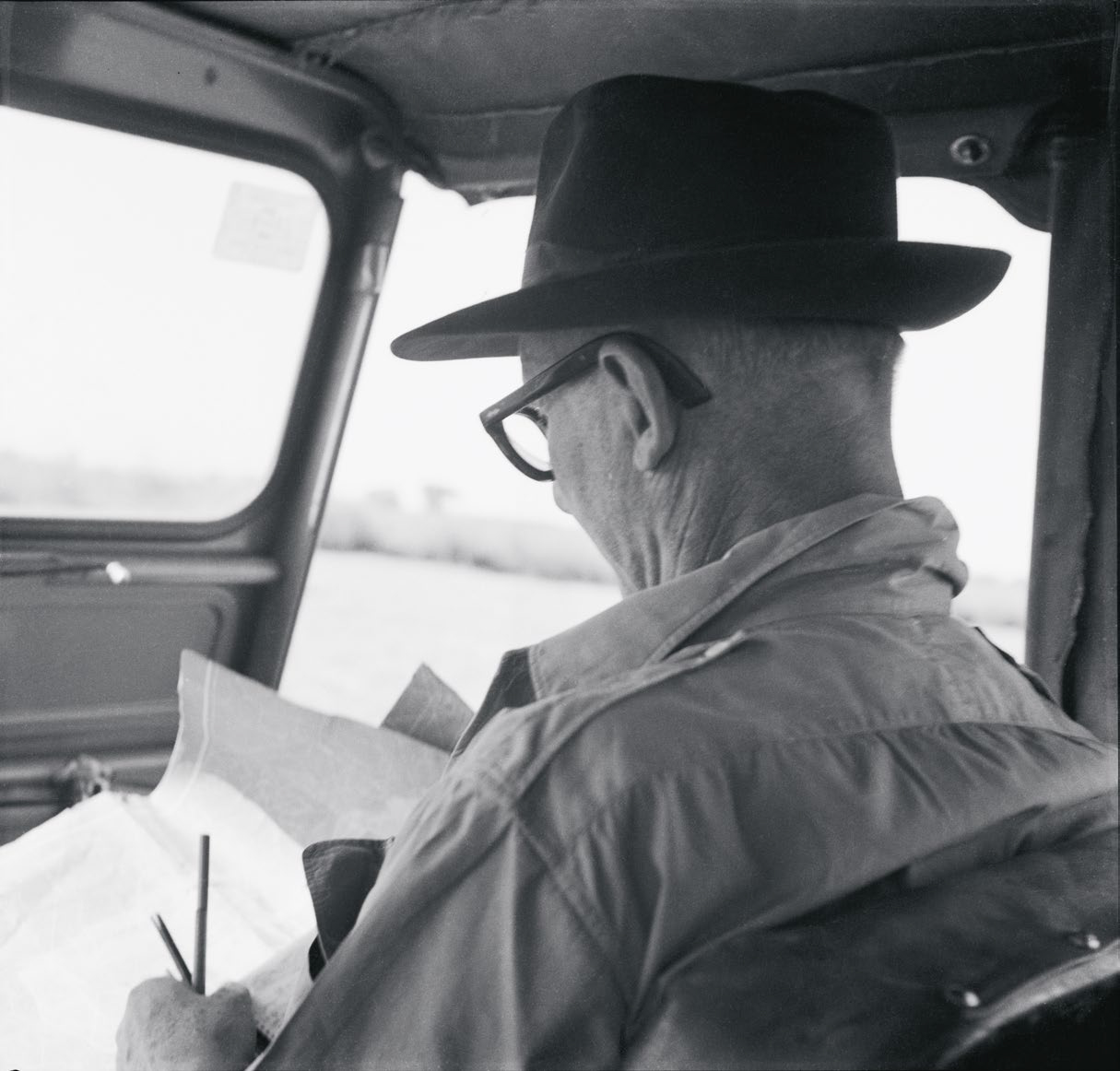 Le Corbusier sitting on the front seat of the jeep while studying a map