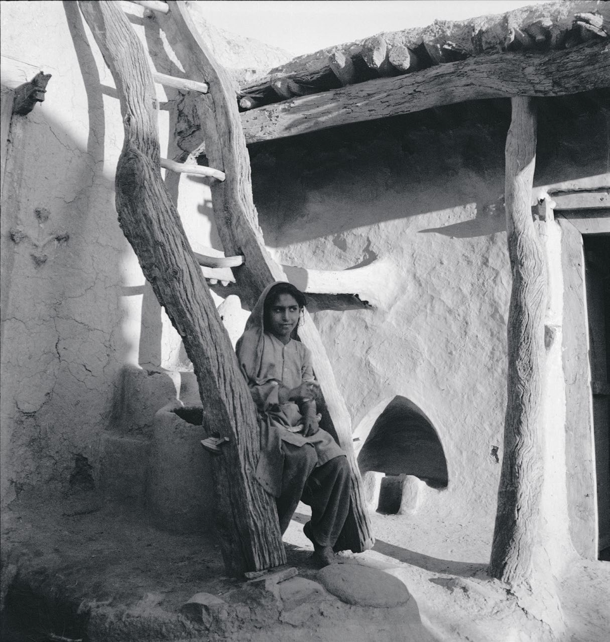 昌迪加尔地区，一名年轻女子坐在走廊外的木梯上。左上角装饰着墙面浮雕和一尊小木雕。