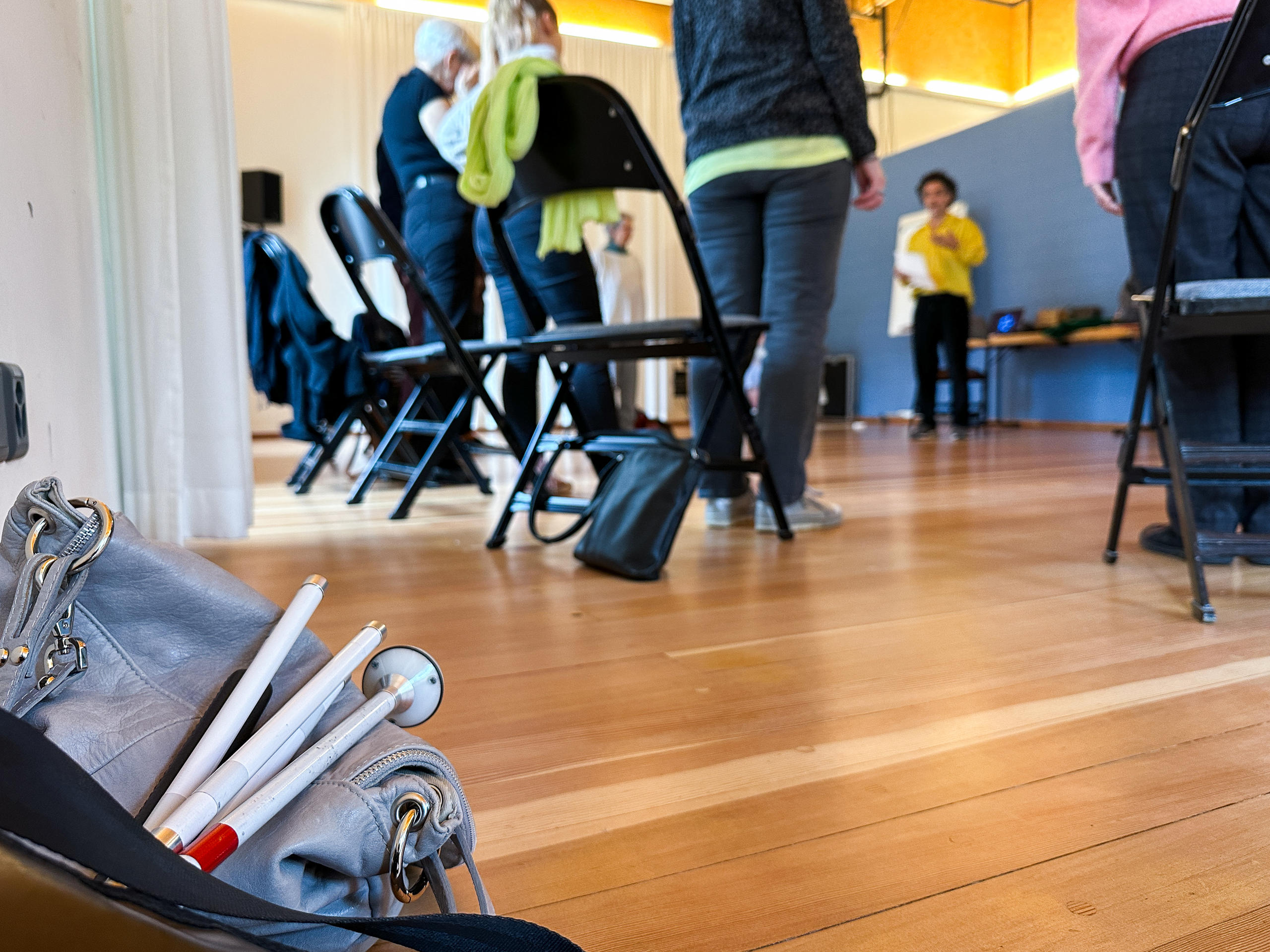 A folded white cane rests on a bag on the floor, in the background there s people standing