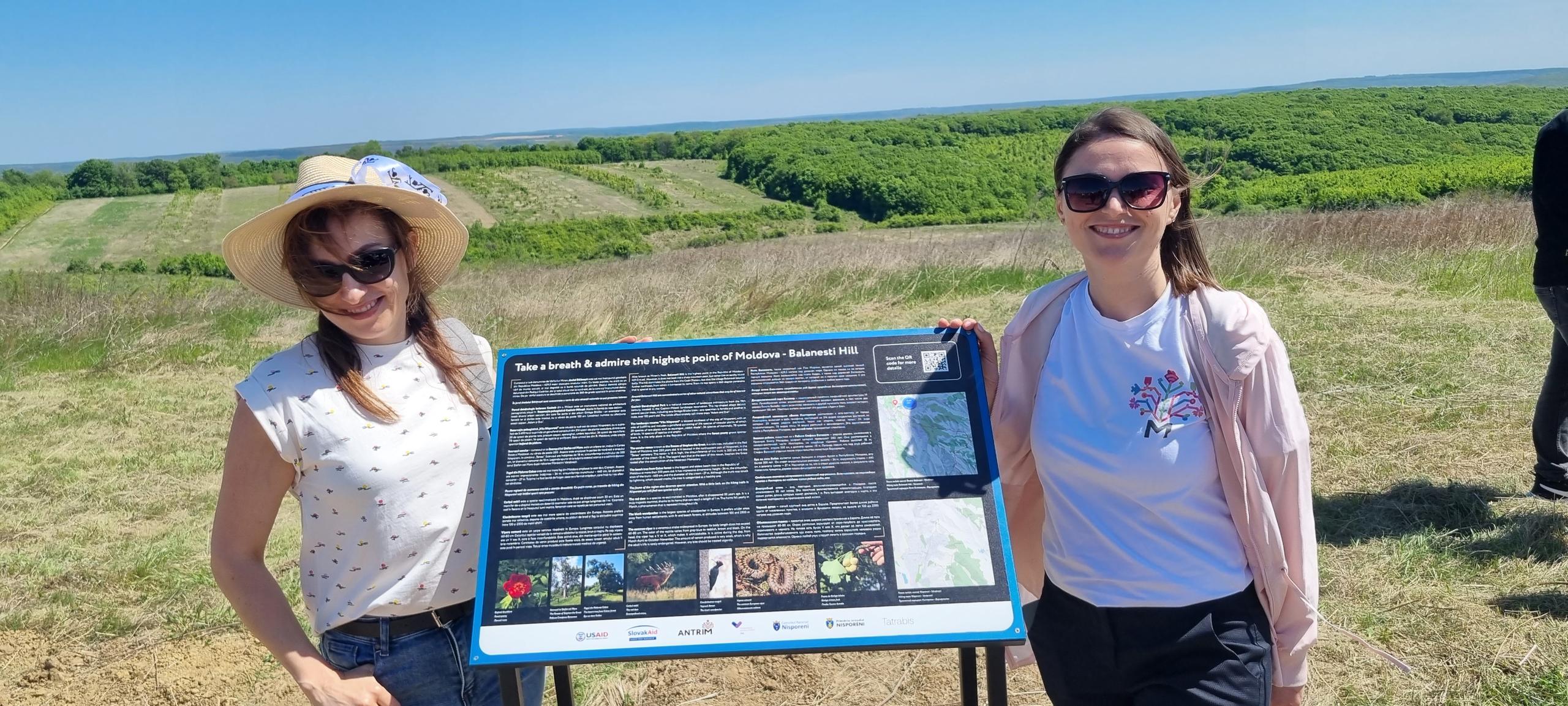 Zwei Frauen neben einem Schild vor einem grünen Feld