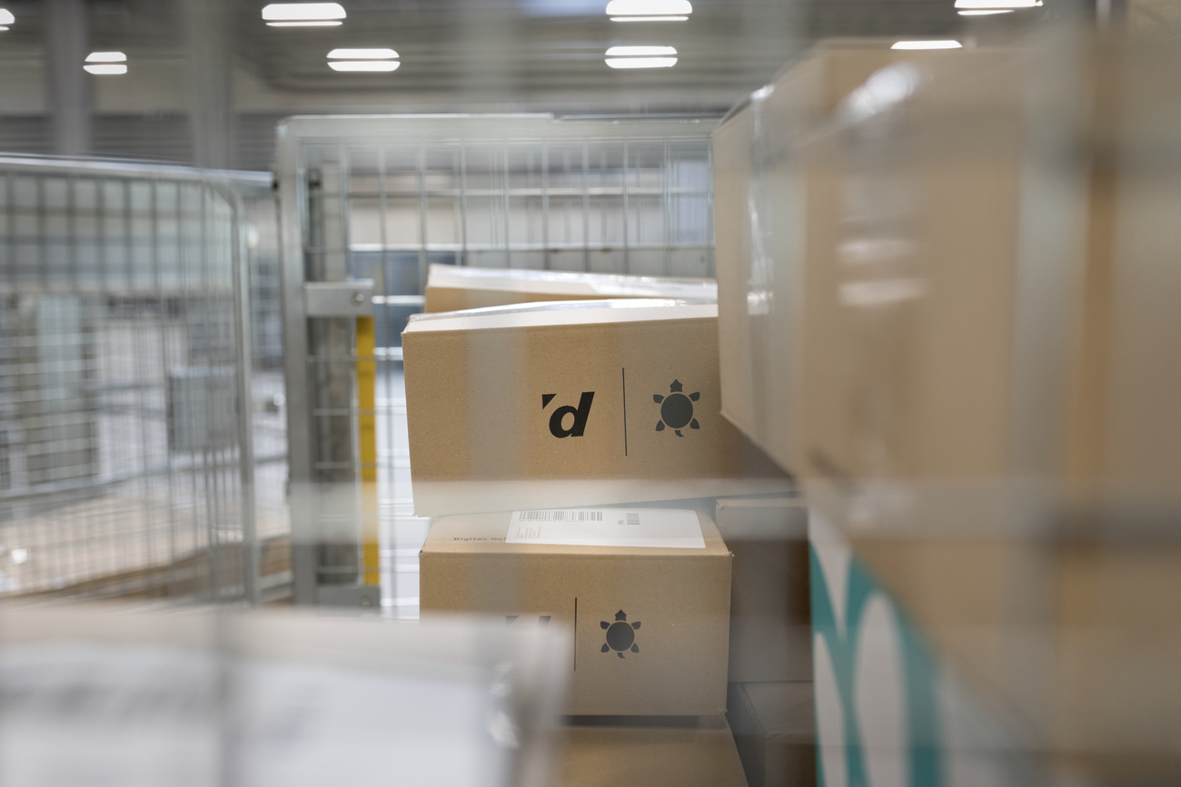 Brown packages piled up in ready for delivery at a warehouse. On the side of the top two packages printed in black ink are the Digitec ‘d’ logo and the Galaxus turtle logo