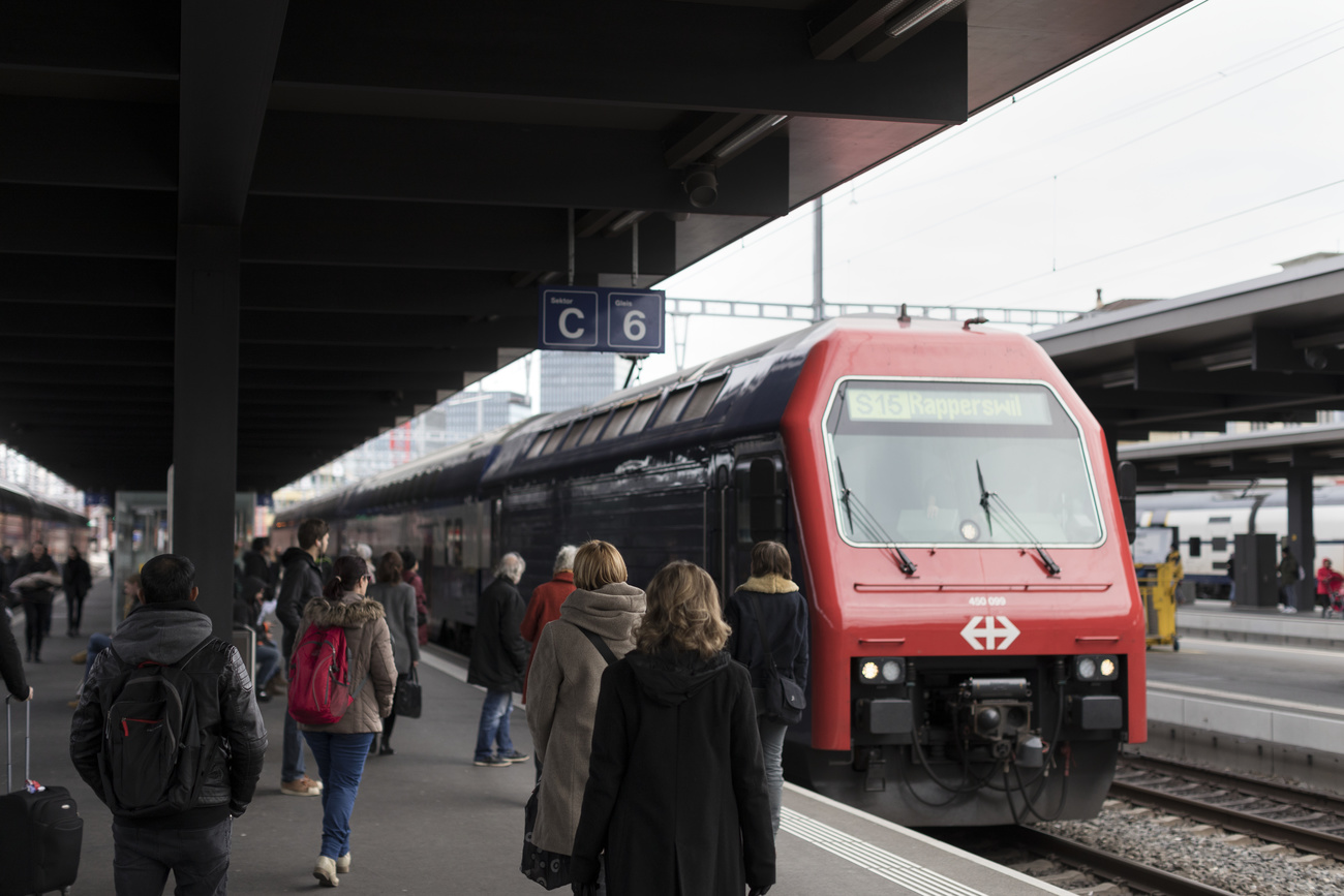 A blue and red S15 commuter train going to Rapperswil arrives at Zurich Oerlikon station. Many passengers are walking towards the train.