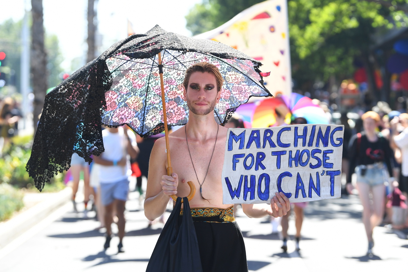 man with makeup carrying black umbrella carrying a sign that say "marching got those who cant"