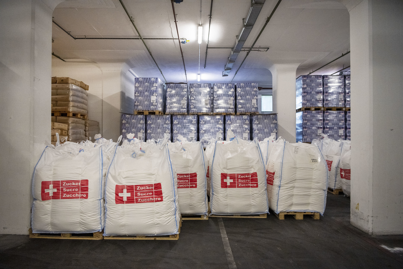 Sugar bags and boxes stored on pallets in the warehouse of the Aarberg sugar factory of Swiss sugar manufacturer Zucker AG. The blue boxes of granulated sugar are towards the back of the photo, while at the front are big white bags printed with the red Zucker AG logo – the Swiss flag (a white plus on a red square background) and next to it says ‘Zucker Sucre Zucchero’