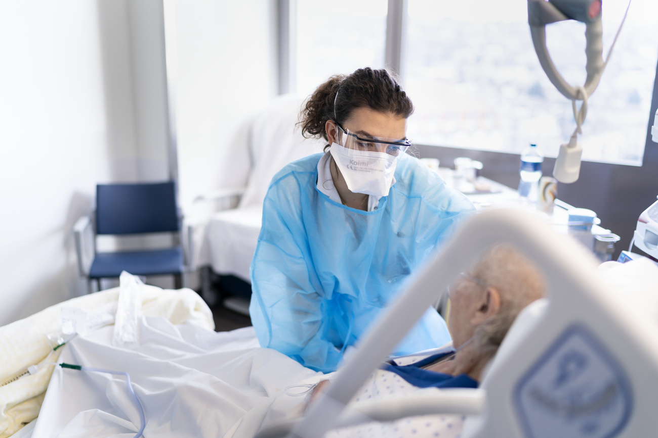 Female doctor wearing a face shield and blue scrubs tending to an elderly man in a hospital bed.