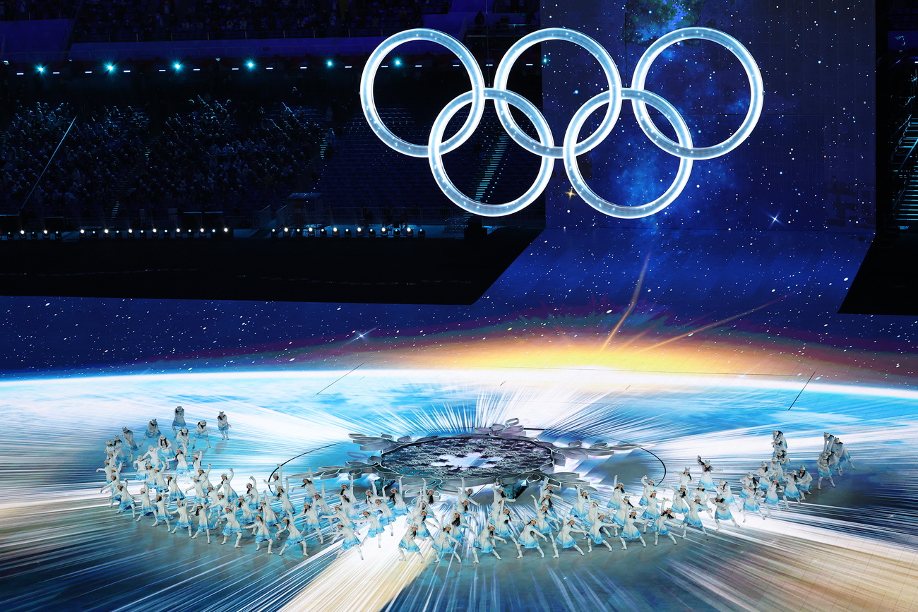 the olympics rings over an ice rink with ice skaters wearing white.