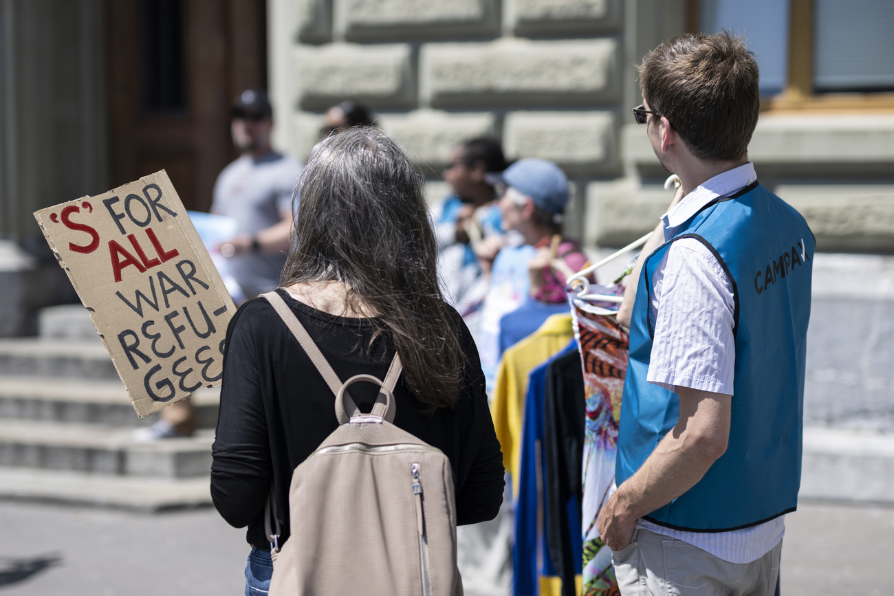 Demonstrierende vor dem Bundeshaus in der Bern mit einem Transparent, auf dem der Schutzstatus S für alle Flüchtenden gefordert wird.