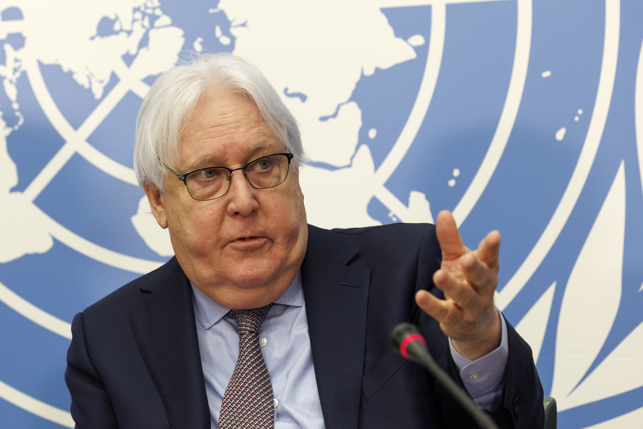 Martin Griffiths, a man with white hair and glasses, is wearing a black suit jacket, blue shirt and black and white checked tie. He sits in front of the blue and white UN flag. The flag features a pair of olive branches and a map of the world. Griffiths’ left hand is raised as he speaks into a microphone in front of him.