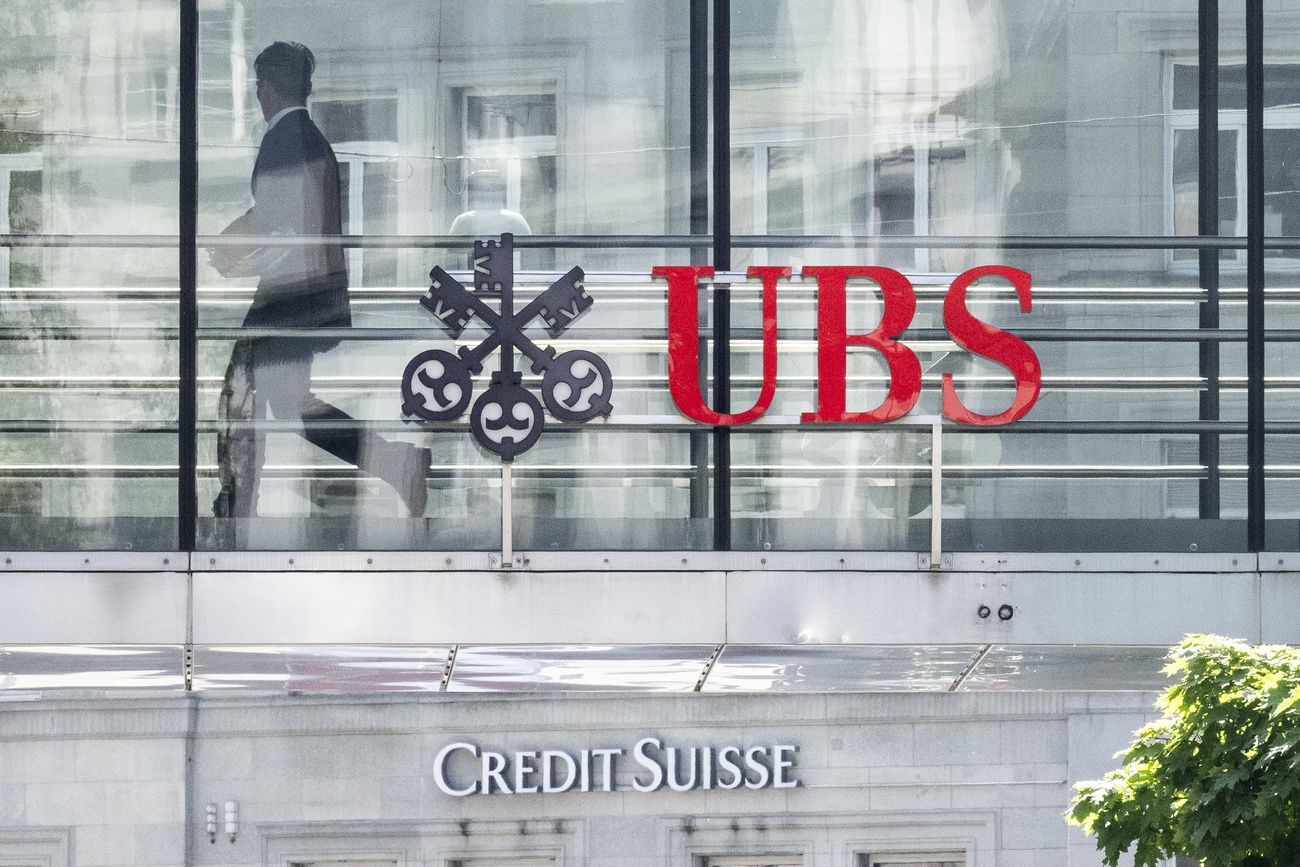 A man can be seen walking behind a glass window. On the outside of the window is the UBS logo, underneath that the Credit Suisse logo