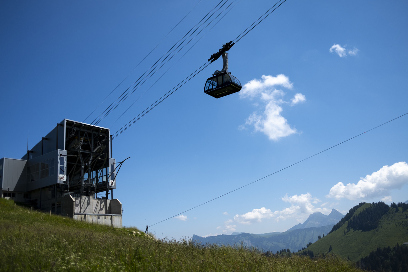 A cable car leaves its station, passing over a green field. It’s a sunny day with blue skies, a few clouds, and Swiss mountains in the background.