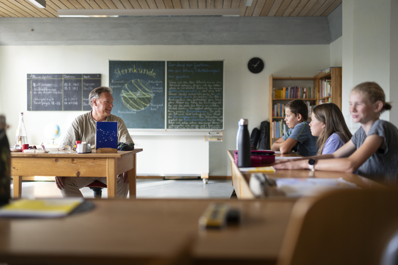 a teacher sits at the front of the classroom. his students sit at their desks in a U-form around him. There is a green chalkboard behind the teacher.