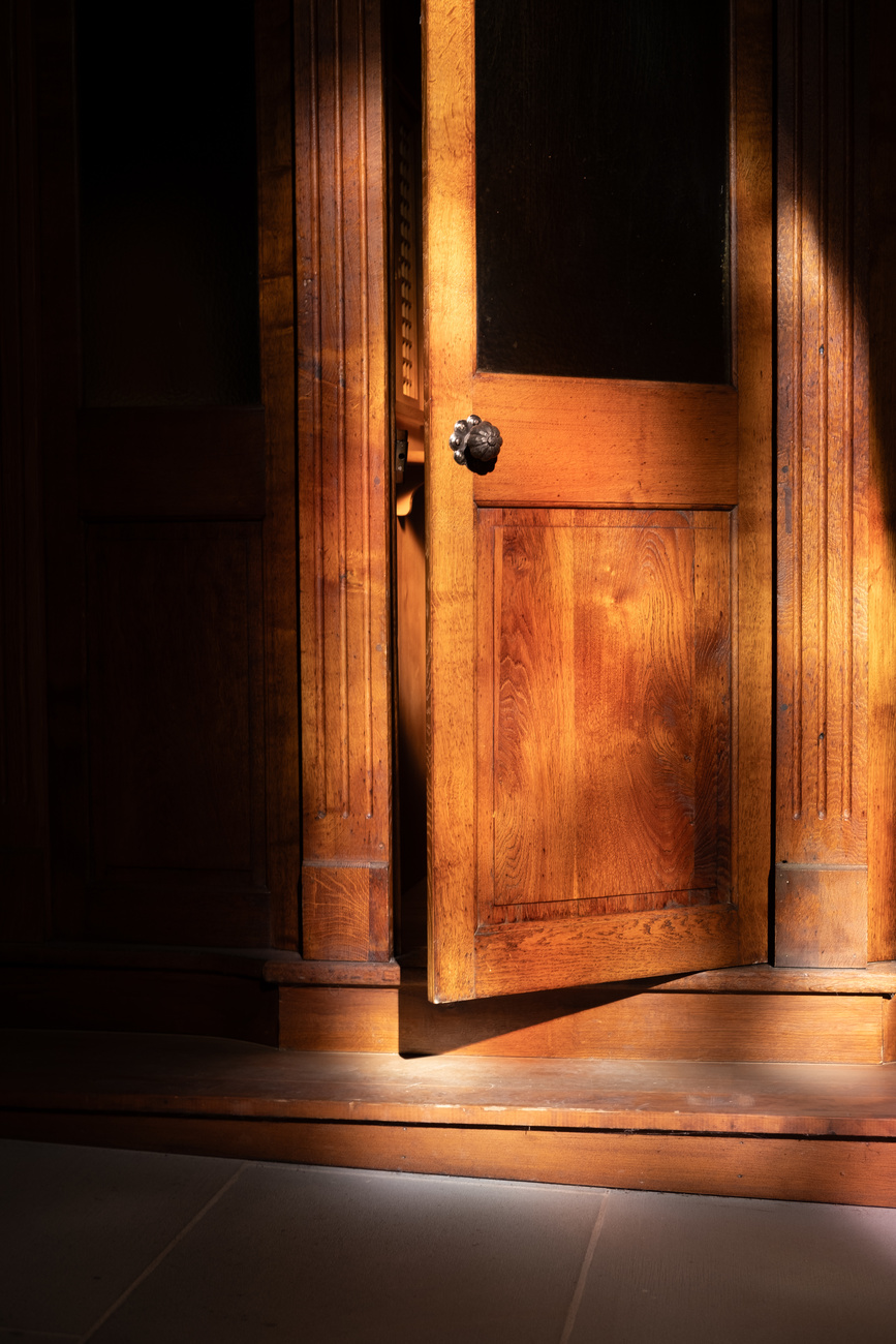 a confessional door in a church is lit up by sun light. The door is open and the confessional box is dark.