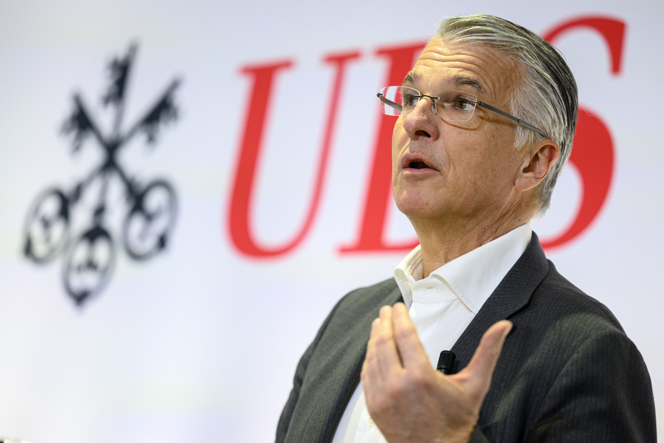 UBS CEO Sergio Ermotti, a man with white hair and glasses, wearing a white shirt and dark jacket, speaks in front of a white background featuring the UBS logo