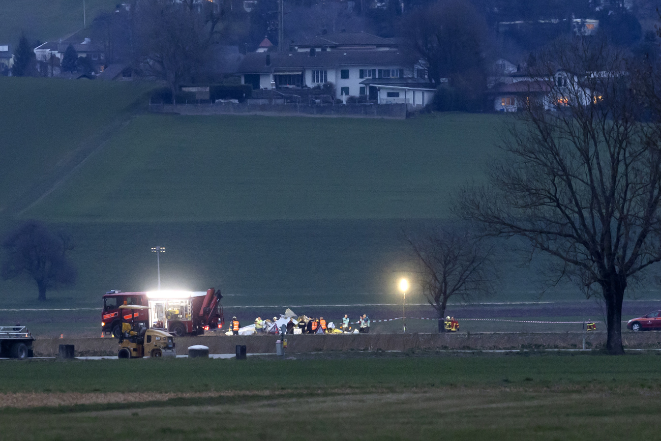 The photo shows a night scene with bright lights of emergency services at Grenchen airstrip, some buildings visible in the background