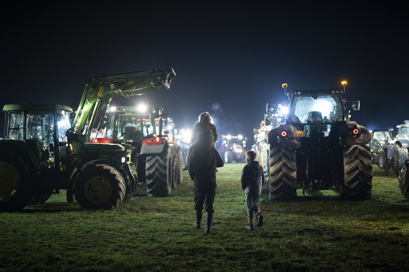 Farming tractors with lights on at night. Mother with two children wearing wellies walk amongst the tractors.