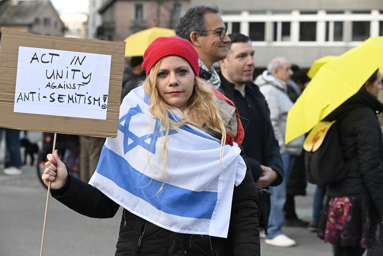 giovane donna bionda a una manifestazione a zurigo, avvolta da una bandiera israeliana, tiene cartello con scritta in inglese "agire uniti contro l'antisemitismo"