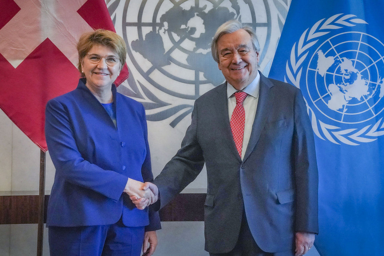 Amherd, left, wearing a blue suit, shakes hands with UN Secretary-General Antonio Guterres wearing a grey suit. Behind them is the red Swiss flag and the blue UN flag