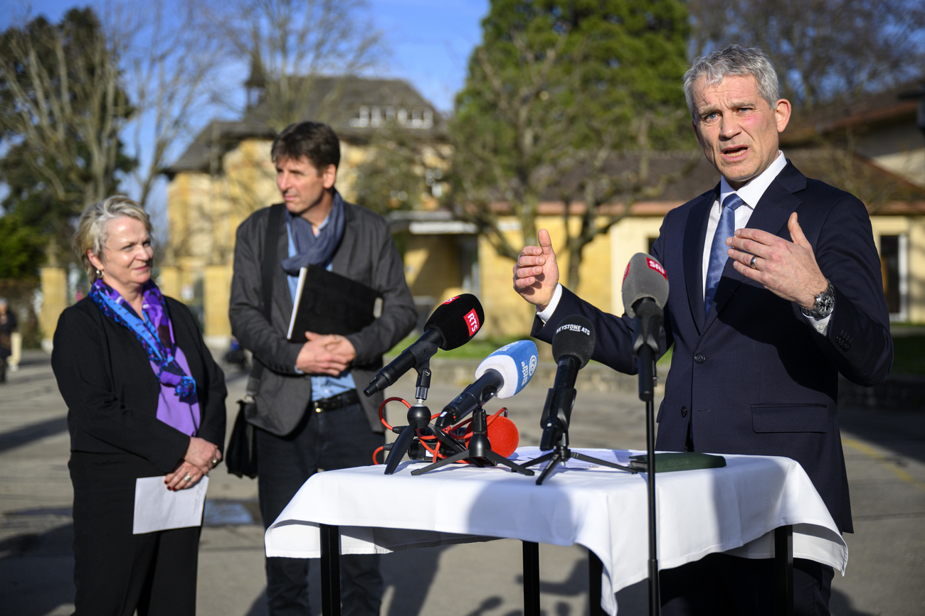 Justice Minister Beat Jans giving a speech. He is wearing a black suit, white shirt and blue tie, and in front of him is a table with a white tablecloth and four media microphones.