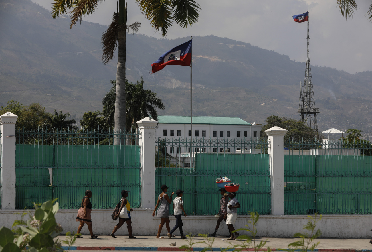 People walk past the National Palace in Port-au-Prince, Haiti, a white building with a turquoise roof with a large white and turquoise wall. The Haiti flag can be seen flying at the centre of the photo with mountains in the background.