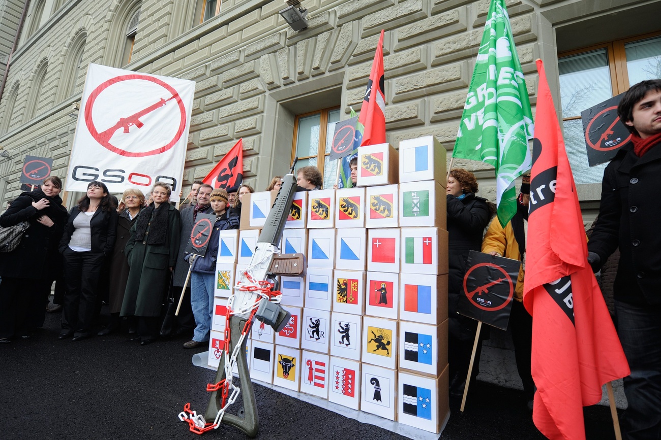 Supporters of the initiative stand in front of the Swiss Federal Palace. A large oversized gun leans on boxes marked with the swiss cantonal flags.