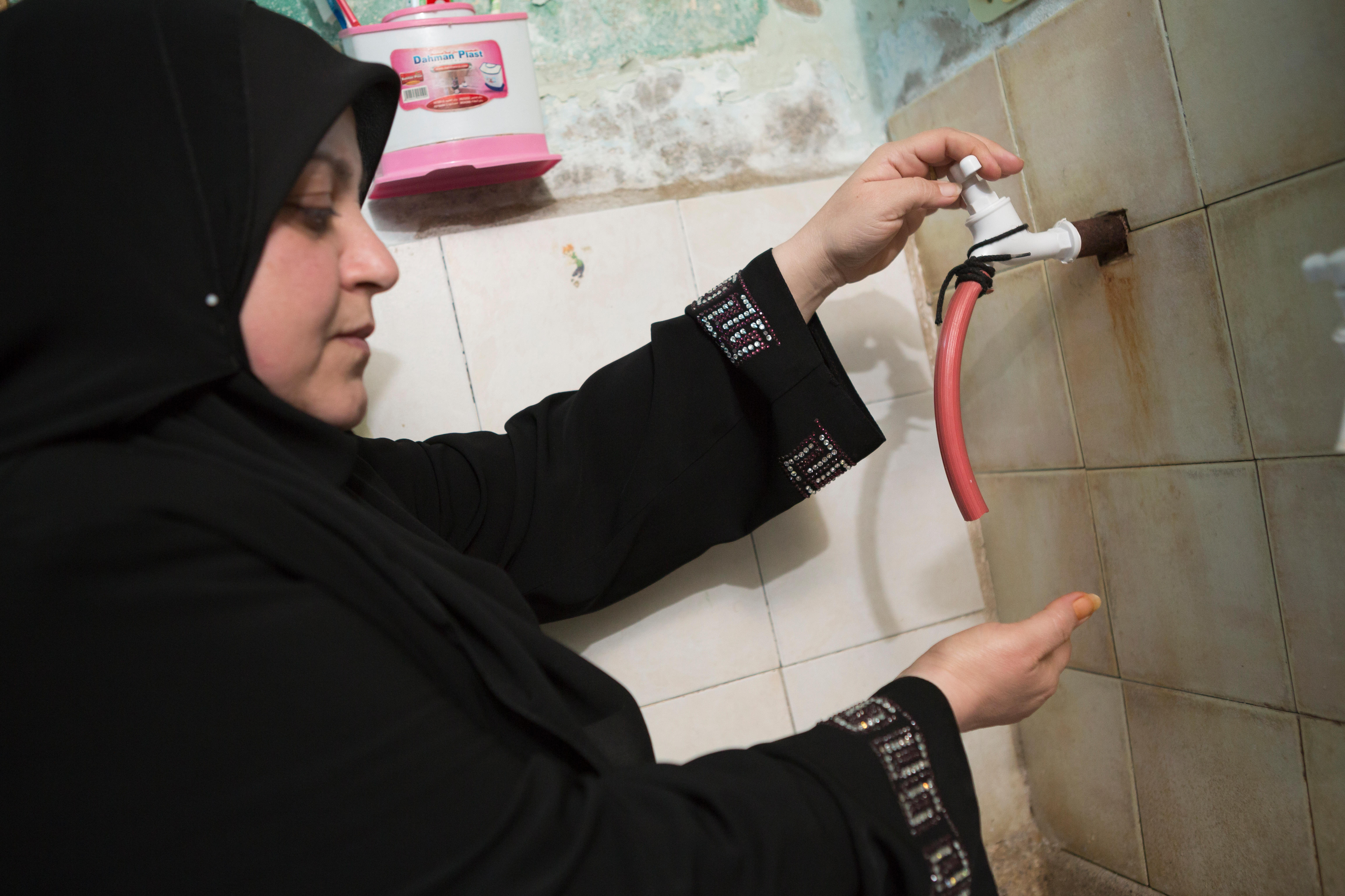 A person with head covering opens a water tap comprises of a short red hose coming out of a tiled wall.