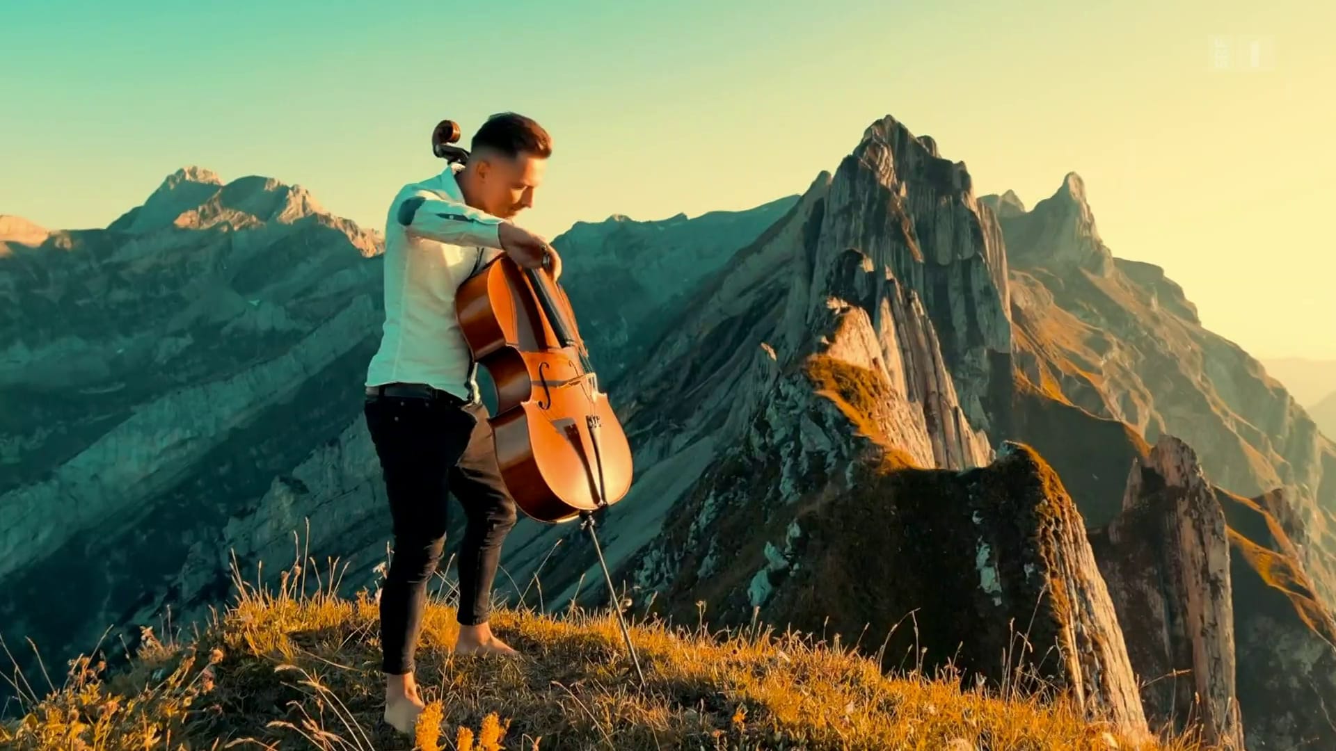 a man in a white button down shirt plays the cello on the top of a mountain with the sun on his face. He is not wearing shoes.